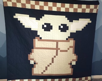 May include: A black and white quilt with a brown and white checkered border. The center of the quilt features a pixelated image of a character from the Star Wars franchise, known as "The Child" or "Baby Yoda".