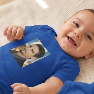 May include: A smiling baby wearing a blue onesie with a rectangular image of a man on the front. The baby is lying on a cream-colored blanket. The onesie has short sleeves.