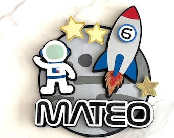 May include: A gray and white cake topper with a rocket, astronaut, and stars. The rocket is red, blue, and white with a number 6 on the side. The astronaut is wearing a white suit with a blue helmet. The stars are yellow. The cake topper says "MATEO" in black letters.