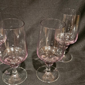 May include: Four stemmed wine glasses with a soft pink hue. The glasses have a rounded bowl and a clear, slender stem. The glasses are arranged on a dark background, showcasing their elegant design.