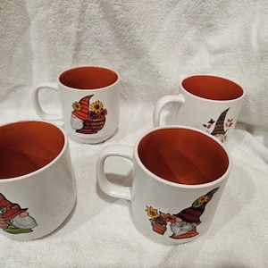 May include: Four white ceramic mugs with orange interiors. Each mug features a colorful gnome design with a hat and flowers. The mugs are ideal for coffee, tea, or other beverages.