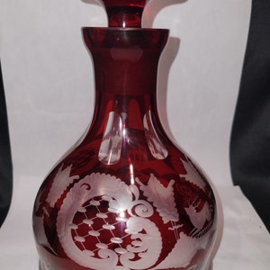May include: A ruby red glass decanter with a matching stopper. The decanter features etched floral and scroll designs. The stopper has a rounded top with circular indentations. The decanter has a bulbous body and a slender neck.