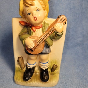 May include: A vintage ceramic figurine of a child playing a banjo. The figurine is painted with a yellow hat, green jacket, blue shorts, and black boots. The child has a red bow tie and is standing on a base with grass and wooden stumps.