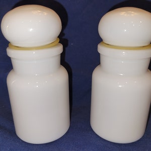 May include: Two white glass apothecary jars with rounded lids. Each jar has a cylindrical body and a slightly flared neck, with a yellow rubber ring between the lid and the jar. The jars are displayed on a blue surface.