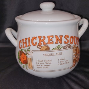 May include: White ceramic soup crock with a lid and two handles. The crock has a colorful illustration of a chicken, vegetables, and spices. The text "CHICKEN SOUP" is printed in orange letters on the front of the crock. The recipe for chicken soup is printed on the side of the crock.