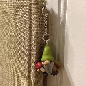 May include: A green and brown gnome figurine with a white beard holding a red and white mushroom. The gnome is attached to a silver chain.