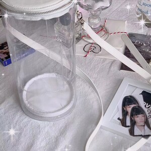May include: A clear, cylindrical handbag with a white top, base, and strap. The bag has a zipper closure and a handle. Other items on a white surface include a framed photo, a glass, and a small gift.