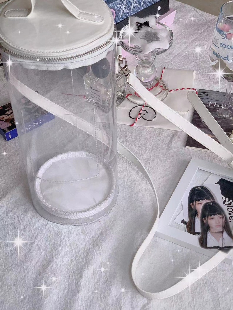 May include: A clear, cylindrical handbag with a white top, base, and strap. The bag has a zipper closure and a handle. Other items on a white surface include a framed photo, a glass, and a small gift.
