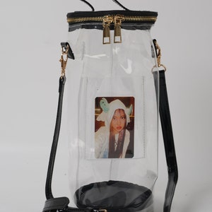 May include: A clear, cylindrical handbag with a black base, trim, and straps. The bag features a gold zipper and hardware, and a small photo is visible inside. The bag has a top handle and a detachable shoulder strap.