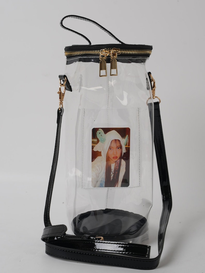 May include: A clear, cylindrical handbag with a black base, trim, and straps. The bag features a gold zipper and hardware, and a small photo is visible inside. The bag has a top handle and a detachable shoulder strap.