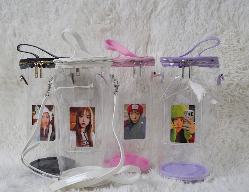 May include: Four clear, rectangular crossbody bags with different colored zippers and handles: black, white, pink, and purple. Each bag has a clear front pocket holding a photo card. The bags are on a white, fluffy surface.