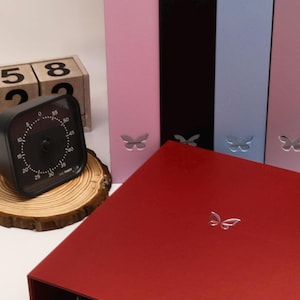 Può includere: Un timer da cucina analogico nero con quadrante bianco è appoggiato su una fetta di legno accanto a un blocchetto di legno con numeri. Una scatola rossa e diverse cartelle colorate con ritagli a farfalla sono sullo sfondo.
