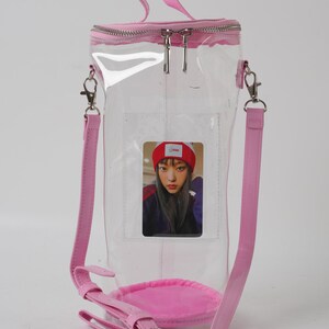 May include: A clear, cylindrical tote bag with a pink zipper, handle, and shoulder strap. The bag features a pink base and a small photo card inside. The bag is designed to be worn over the shoulder or carried by the handle.
