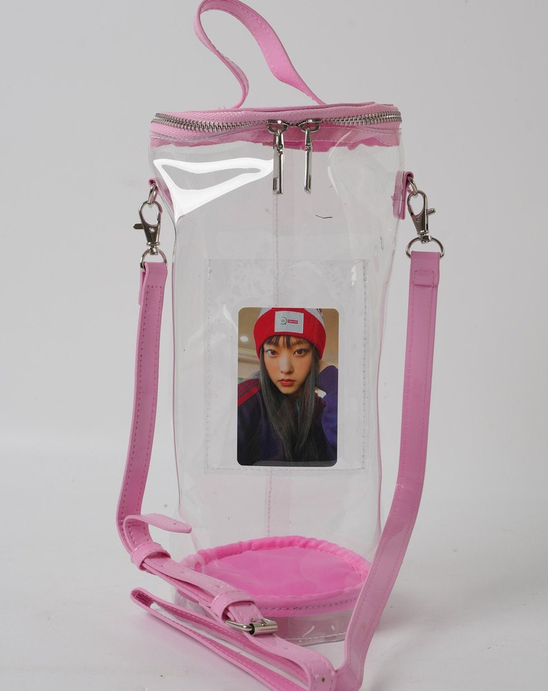 May include: A clear, cylindrical tote bag with a pink zipper, handle, and shoulder strap. The bag features a pink base and a small photo card inside. The bag is designed to be worn over the shoulder or carried by the handle.