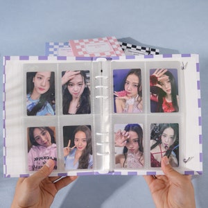 May include: A purple and white checkered binder with clear plastic sleeves holding photo cards of a K-pop group. The binder has a metal ring binding system.