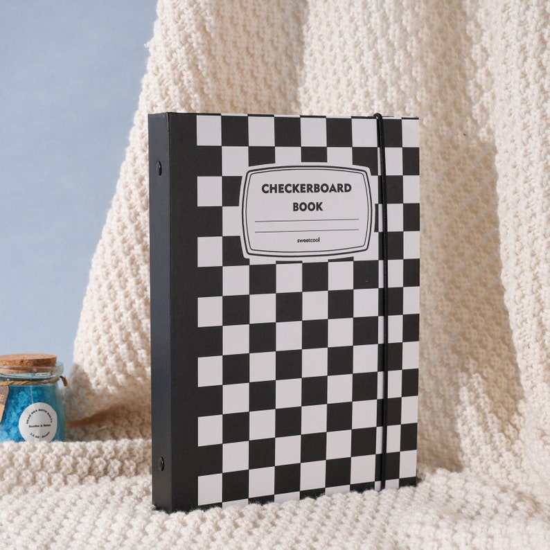 May include: Black and white checkered notebook with a black spine and a black elastic band closure. The cover has the text "Checkerboard Book" in white lettering.