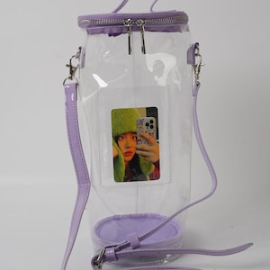 May include: A clear, cylindrical handbag with a lavender zipper, handle, and strap. The bag features a small, rectangular pocket with a photo of a person wearing a green hat. The bag has a lavender base.