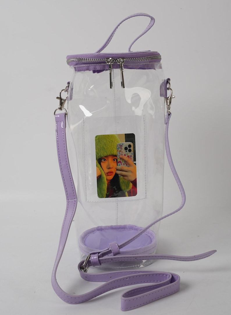 May include: A clear, cylindrical handbag with a lavender zipper, handle, and strap. The bag features a small, rectangular pocket with a photo of a person wearing a green hat. The bag has a lavender base.