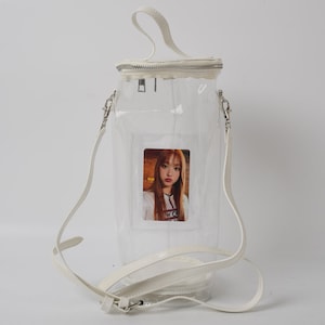 May include: Clear, cylindrical handbag with a white handle and shoulder strap. The bag has a zipper closure and a small photo of a person inside. The bag is made of clear plastic and has white trim.
