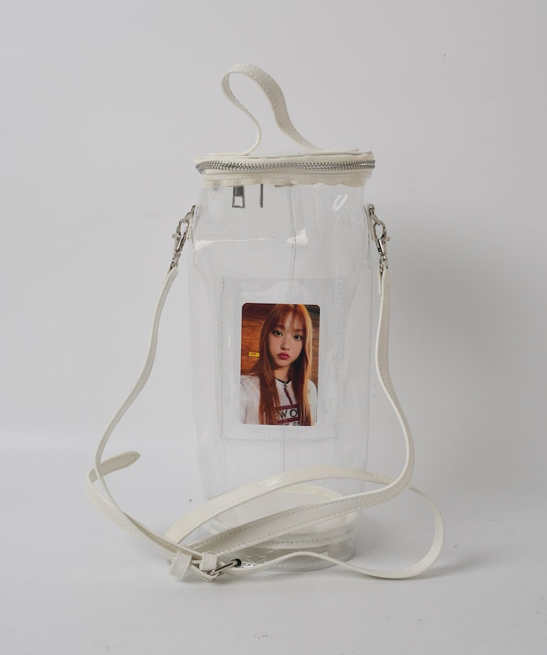 May include: Clear, cylindrical handbag with a white handle and shoulder strap. The bag has a zipper closure and a small photo of a person inside. The bag is made of clear plastic and has white trim.