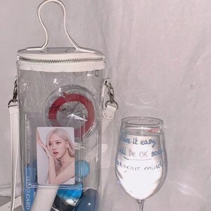May include: A clear, cylindrical handbag with a white handle and strap, filled with various items including a photo card and blue spheres. A wine glass with handwritten text that reads "Take it easy, all will be OK soon, relax your mind" is next to the bag.