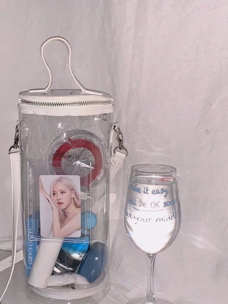 May include: A clear, cylindrical handbag with a white handle and strap, filled with various items including a photo card and blue spheres. A wine glass with handwritten text that reads "Take it easy, all will be OK soon, relax your mind" is next to the bag.