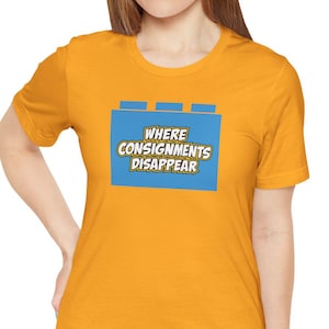 May include: A mustard yellow short-sleeve t-shirt featuring a blue rectangular graphic shaped like a building block. The graphic contains the text WHERE CONSIGNMENTS DISAPPEAR in white letters with a dark outline, centred on the chest.
