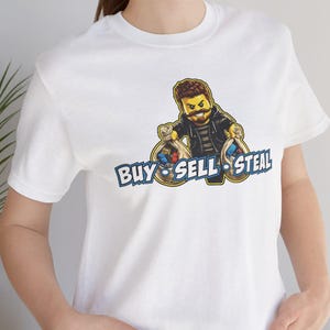 May include: A white short-sleeved t-shirt featuring a graphic of a cartoon character with a beard and dark clothing holding two money bags. Below the character, the text reads "BUY SELL STEAL" in bold blue letters with a white outline and black shadow.