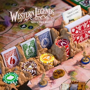 Peut inclure: Jeu de société "Western Legends Mountain Range" avec des cartes, des pions et des jetons de poker. Les cartes présentent divers motifs et les jetons sont de différentes couleurs. Le plateau de jeu est beige et marron.