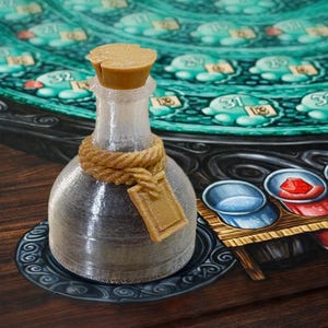 Puede incluir: Una botella de vidrio transparente con un tapón de corcho y una cuerda atada alrededor del cuello. Una pequeña etiqueta cuelga de la cuerda. La botella se encuentra sobre un plato decorativo, con un tablero de juego en el fondo.