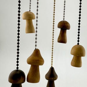 May include: A collection of wooden mushroom-shaped light pull chains in varying shades of brown. Each mushroom is suspended by a beaded chain, with colors including black, silver, and gold. The mushrooms have a natural, organic aesthetic.