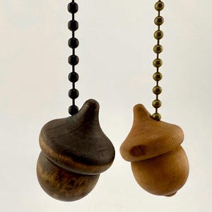 May include: Two wooden acorn-shaped ceiling fan pull chains. One is dark brown and attached to a black beaded chain. The other is light brown and attached to a brass-colored beaded chain. The acorns have a natural wood grain.