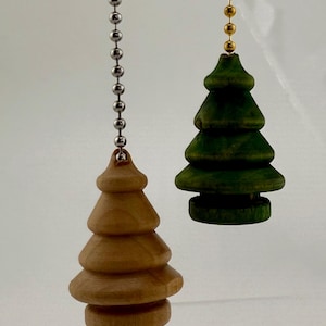 May include: Two Christmas tree-shaped light pull chains. One is natural wood-toned, the other is green. Both are suspended by a metal ball chain. The tree shapes have a tiered design, and are likely made of wood.