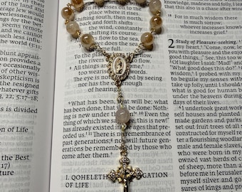 Può includere: Un rosario con perline color oro e un ciondolo a forma di croce dorata. Il rosario è appoggiato su un libro aperto con testo. Le perline sono di colore marrone chiaro.