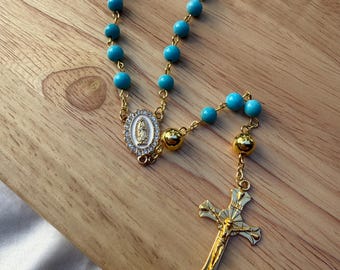 Può includere: Un rosario dorato con perline turchesi. Il rosario presenta una croce dorata con una raffigurazione di Gesù e un medaglione dorato con un'immagine religiosa. Il rosario è esposto su una superficie di legno.