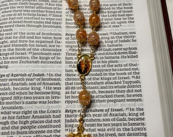 Può includere: Un rosario con perline color ambra e una croce dorata, appoggiato su un libro aperto. Le perline sono intervallate da elementi decorativi dorati e una piccola immagine.
