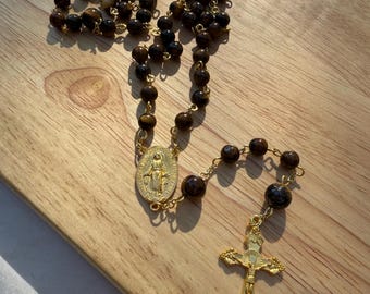 Può includere: Un rosario con perline marroni occhio di tigre e dettagli in metallo color oro. Il rosario presenta un crocifisso color oro e un ciondolo ovale color oro con l'immagine della Vergine Maria.