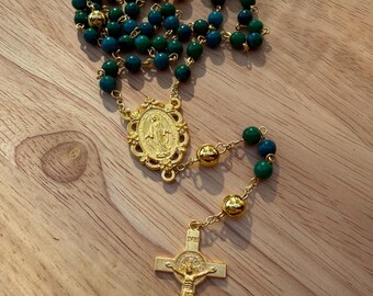 Può includere: Rosario dorato con perline blu e verdi. Il rosario presenta un crocifisso dorato con una figura di Gesù e un ciondolo dorato con un'immagine religiosa. Le perline sono infilate su una catena d'oro.