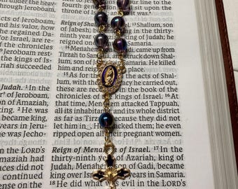 Può includere: Un rosario con perline viola scuro e dettagli in metallo color oro. Il rosario presenta un ciondolo a croce e un medaglione con un'immagine religiosa. Il rosario è appoggiato su un libro aperto.