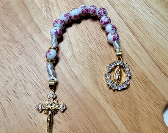 Può includere: Un rosario con perline bianche decorate con motivi floreali rosa. Il rosario presenta un ciondolo a forma di crocifisso dorato e un ciondolo con una raffigurazione della Vergine Maria, circondata da cristalli trasparenti.