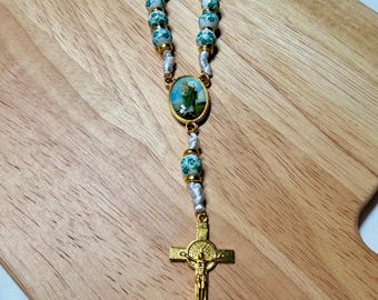 Può includere: Un rosario con perline bianche e verdi, dettagli in metallo color oro e un crocifisso dorato. Il centro presenta una piccola immagine ovale di una figura religiosa. Le perline hanno un motivo floreale.