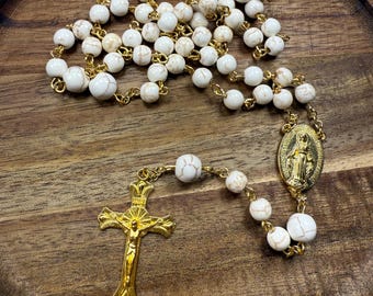 Può includere: Un rosario con perline bianche e macchiate di marrone infilate su una catena color oro. Alla fine del rosario ci sono un crocifisso color oro e un medaglione color oro con una figura religiosa. Il rosario è esposto su un vassoio di legno.