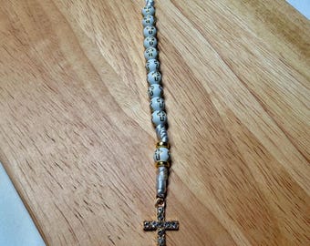 Può includere: Un rosario con perline bianche, ognuna ornata da una croce, infilate su un cordoncino d'argento. Il rosario presenta un ciondolo a forma di croce dorata con cristalli incastonati e un elemento superiore decorativo.