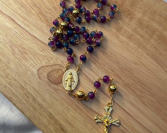 Può includere: Un rosario con perline viola e blu, catena color oro e un crocifisso dorato. Le perline sono intervallate da perline color oro. Il rosario presenta anche un ciondolo ovale color oro con una figura religiosa.