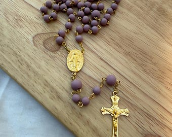 Può includere: Un rosario con perline viola opache e dettagli in metallo color oro. Il rosario presenta un crocifisso e un medaglione con una figura religiosa. Le perline sono infilate su una catena d'oro, presentato su una superficie di legno.