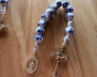 Può includere: Un rosario con perline bianche e blu a motivi floreali. Il rosario presenta un crocifisso color oro e un ciondolo color oro con una figura. Il rosario è su una superficie di legno.