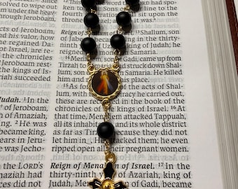 Può includere: Un rosario con perline nere opache e un ciondolo a forma di croce color oro. La croce presenta dettagli in smalto nero. È presente anche un piccolo ciondolo ovale con un'immagine religiosa.