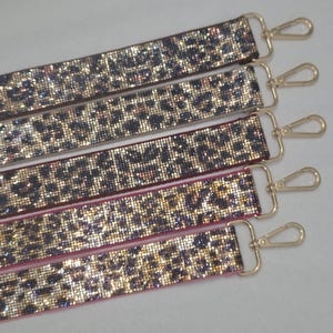 May include: Five rectangular bag straps featuring a dense, sparkling leopard print pattern in shades of gold, black, and brown. Each strap is finished with a polished gold-tone metal lobster clasp attached to a rectangular loop at one end.