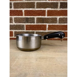 May include: A small, stainless steel saucepan with a black handle and a metal loop for hanging. The pan has a rounded base and a shiny, metallic finish. The handle is attached with two rivets.