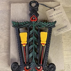 Puede incluir: Un calientaplatos de hierro fundido negro con un diseño verde y rojo. El calientaplatos presenta dos escobas amarillas y el texto "Made in America" y "John Wright".